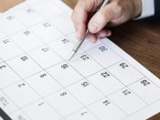 Mão de homem segurando caneta prateada e apontando para o dia 16 em um calendário de mesa branco, representando o planejamento de feriados em Tubarão 2026.