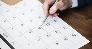 Mão de homem segurando caneta prateada e apontando para o dia 16 em um calendário de mesa branco, representando o planejamento de feriados em Tubarão 2026.