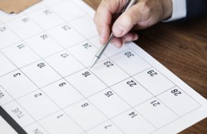 Mão de homem segurando caneta prateada e apontando para o dia 16 em um calendário de mesa branco, representando o planejamento de feriados em Tubarão 2026.