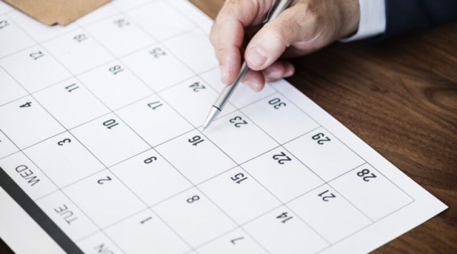 Businessman marking on calendar for an appointment Mão de homem segurando caneta prateada e apontando para o dia 16 em um calendário de mesa branco, representando o planejamento de feriados em Tubarão 2026.