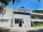 Fachada da Câmara de Vereadores de Tubarão em um dia ensolarado. O edifício é uma construção de alvenaria branca com dois pavimentos. Na parte central, acima da porta de vidro da recepção, destaca-se o letreiro prateado com o nome da instituição. À direita, há uma área gramada com plantas ornamentais e uma rampa de acesso. Árvores emolduram a lateral esquerda da foto sob um céu azul claro.