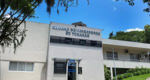Fachada da Câmara de Vereadores de Tubarão em um dia ensolarado. O edifício é uma construção de alvenaria branca com dois pavimentos. Na parte central, acima da porta de vidro da recepção, destaca-se o letreiro prateado com o nome da instituição. À direita, há uma área gramada com plantas ornamentais e uma rampa de acesso. Árvores emolduram a lateral esquerda da foto sob um céu azul claro.