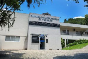 Fachada da Câmara de Vereadores de Tubarão em um dia ensolarado. O edifício é uma construção de alvenaria branca com dois pavimentos. Na parte central, acima da porta de vidro da recepção, destaca-se o letreiro prateado com o nome da instituição. À direita, há uma área gramada com plantas ornamentais e uma rampa de acesso. Árvores emolduram a lateral esquerda da foto sob um céu azul claro.
