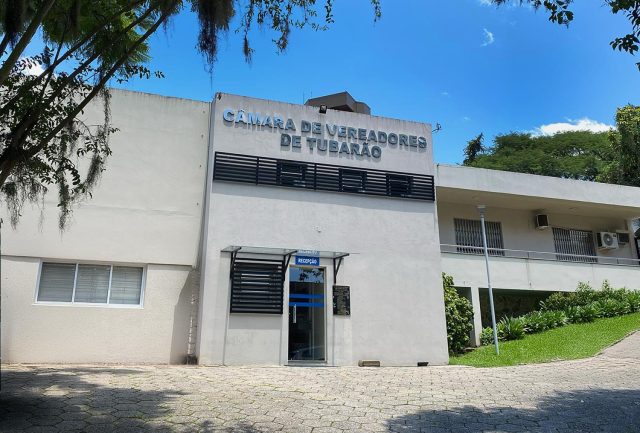Fachada da Câmara de Vereadores de Tubarão em um dia ensolarado. O edifício é uma construção de alvenaria branca com dois pavimentos. Na parte central, acima da porta de vidro da recepção, destaca-se o letreiro prateado com o nome da instituição. À direita, há uma área gramada com plantas ornamentais e uma rampa de acesso. Árvores emolduram a lateral esquerda da foto sob um céu azul claro.