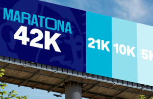 Outdoor publicitário da Maratona de Tubarão 2026 exibindo as distâncias de 42K, 21K, 10K e 5K em diferentes tons de azul. No canto direito, o logotipo do evento com a data 24 de maio de 2026 sobre um fundo de céu azul e árvores.