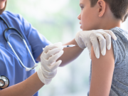 Fotografia em plano médio mostrando uma profissional de saúde, vestindo jaleco azul e estetoscópio, aplicando uma vacina no braço de um adolescente de aproximadamente 12 anos. O jovem está sentado de perfil, vestindo uma camiseta cinza, enquanto a enfermeira utiliza luvas brancas e uma seringa para realizar o procedimento em um ambiente clínico iluminado.