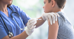 Fotografia em plano médio mostrando uma profissional de saúde, vestindo jaleco azul e estetoscópio, aplicando uma vacina no braço de um adolescente de aproximadamente 12 anos. O jovem está sentado de perfil, vestindo uma camiseta cinza, enquanto a enfermeira utiliza luvas brancas e uma seringa para realizar o procedimento em um ambiente clínico iluminado.