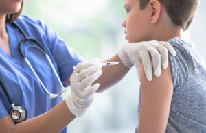 Fotografia em plano médio mostrando uma profissional de saúde, vestindo jaleco azul e estetoscópio, aplicando uma vacina no braço de um adolescente de aproximadamente 12 anos. O jovem está sentado de perfil, vestindo uma camiseta cinza, enquanto a enfermeira utiliza luvas brancas e uma seringa para realizar o procedimento em um ambiente clínico iluminado.