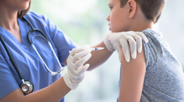 Fotografia em plano médio mostrando uma profissional de saúde, vestindo jaleco azul e estetoscópio, aplicando uma vacina no braço de um adolescente de aproximadamente 12 anos. O jovem está sentado de perfil, vestindo uma camiseta cinza, enquanto a enfermeira utiliza luvas brancas e uma seringa para realizar o procedimento em um ambiente clínico iluminado.